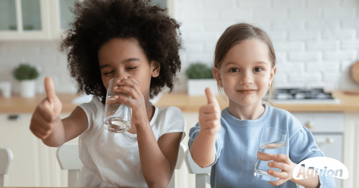 Ein Mädchen mit dunkler Haut und einem Afro und ein Mädchen mit weißer Haut und einem Zopf sind in einer Küche mit hellen Bauernmöbel. Das Mädchen mit weißer Haut schaut in die Kamera und hält in der einen Hand ein Wasserglas und mit der anderen Hand den Daumen nach oben. Das andere Mädchen ist damit beschäftig aus dem Glas zu trinken und hält auch den Daumen nach oben.