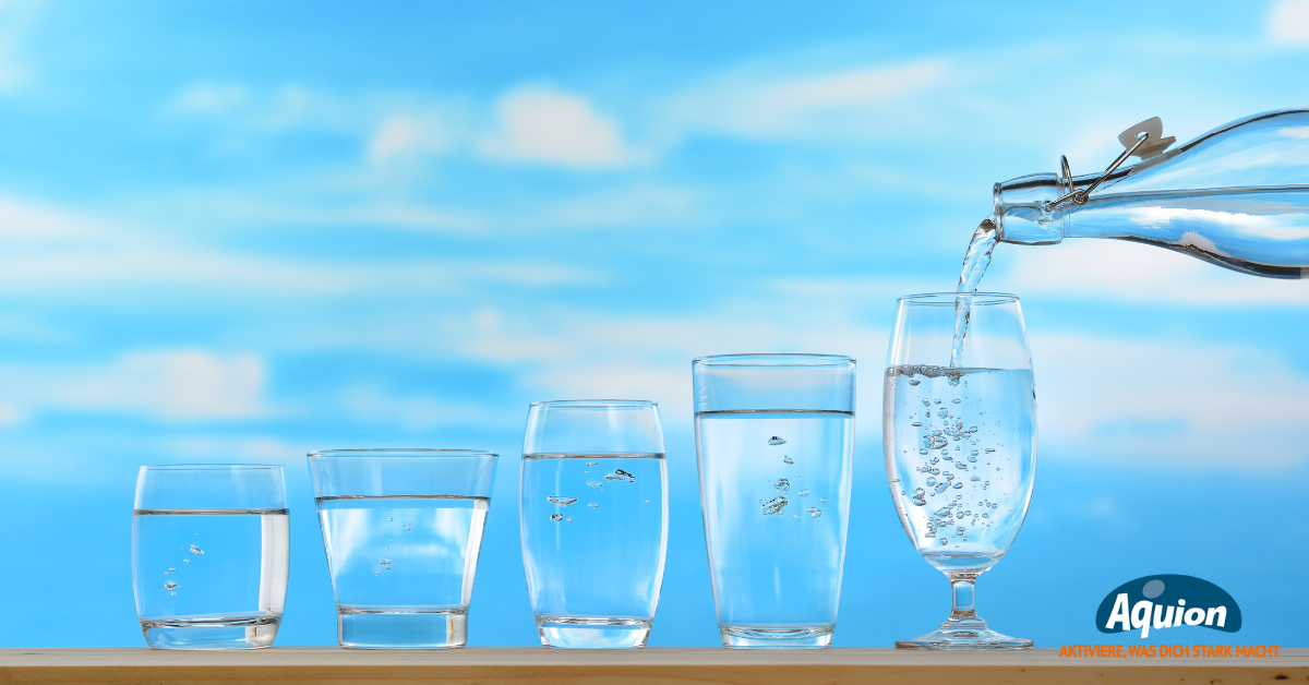 Auf einem Brett stehen 5 unterschiedlich große Gläser von schlicht zu schön, von klein zu groß. In das letzte Glas wird aus einer Glasflasche Trinkwasser geschüttet.Im Hintergrund ein blauer Himmel mit Schleier