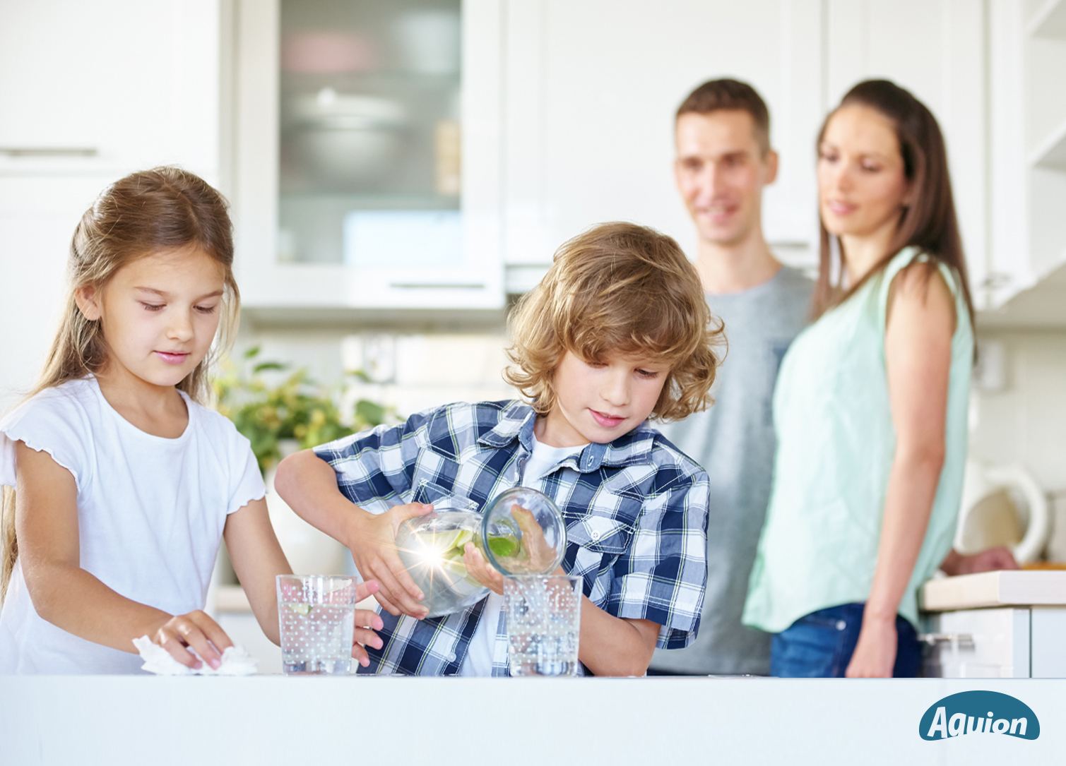 Ein Junge gießt Wasser aus einer Karaffe in ein Glas. Seine Schwester steht neben ihm und schaut zu. Die Eltern stehen im Hintergrund und schauen auf die Kinder. 