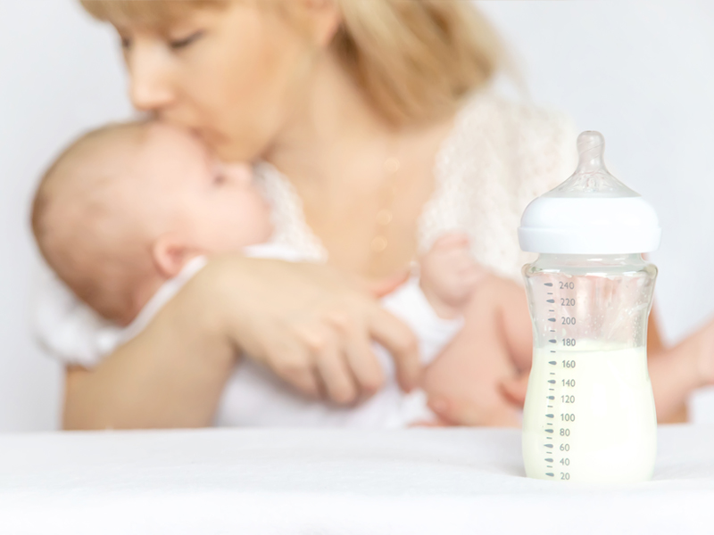 Eine blonde Frau hält ein Baby im Arm und küsst es. Im Vordergrund des Bildes steht eine Babyflasche, die zur Hälfte mit flüssiger Babynahrung gefüllt ist. 