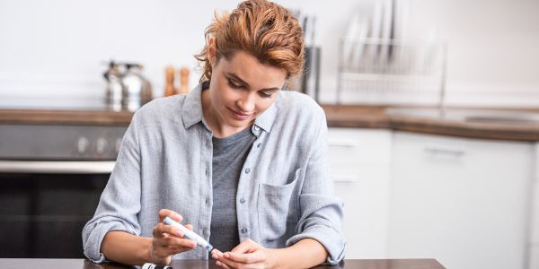 Eine junge weiße Frau mit rotblonden Haaren, sitzt in einer Küche an einem Küchentisch und pickst sich mit einem Pen in den Finger, um ihren Blutzucker zu analysieren.