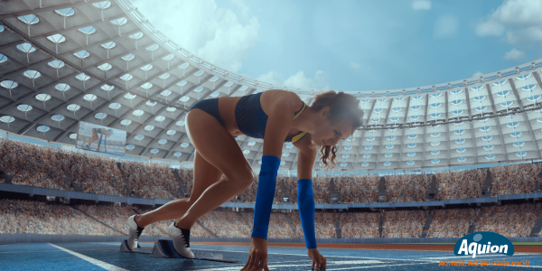 Eine Läuferin in einem blauen Laufset ist in einem voll besetzten Sportstadion und befindet sich in der Startposition, um mit dem Rennen zu beginnen. Sie hat sich auf ihren Fingern abgestützt und die Füße auf die Starthilfe gestellt.
