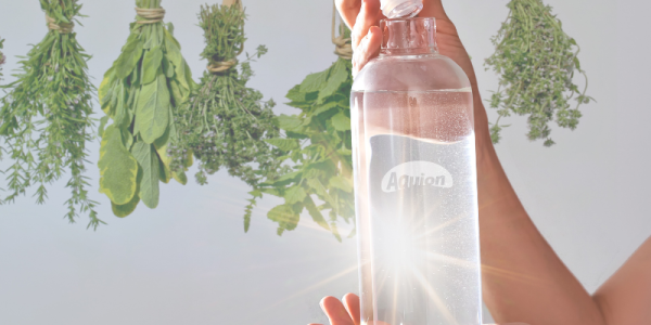 Eine Flasche Wasser wird in der Hand gehalten. Hinten im Bild sind getrocknete Kräuter in Büschel zu sehen, die zu den Bitterstoffen gehören.