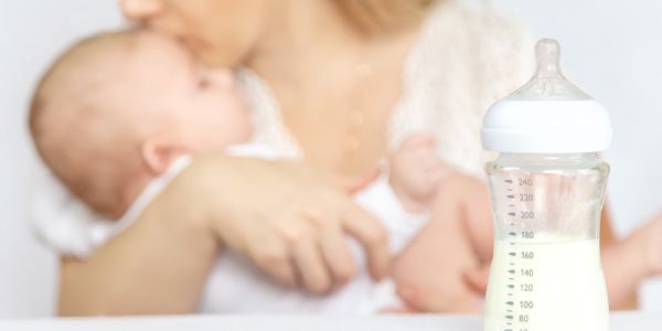 Eine blonde Frau hält ein Baby im Arm und küsst es. Im Vordergrund des Bildes steht eine Babyflasche, die zur Hälfte mit flüssiger Babynahrung gefüllt ist.