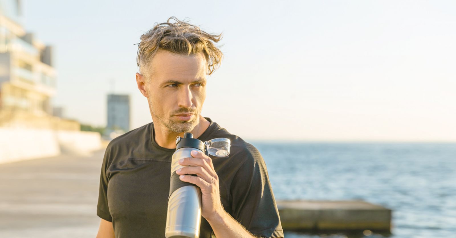 Mann steht am Hafen und hält eine Sporttrinkflasche in der Hand. Er trägt ein schwarzes T-Shirt und blickt nach links.